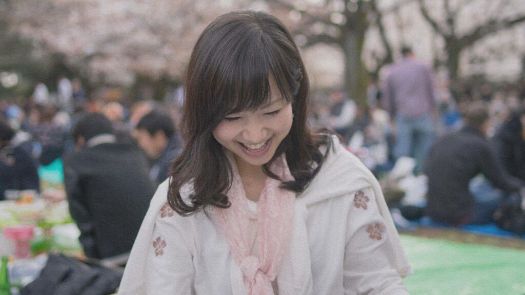 Jeune femme japonaise souriant lors d'un pique-nique sous les cerisiers en fleurs pendant le Hanami, symbole de partage et de célébration de l’éphémère.