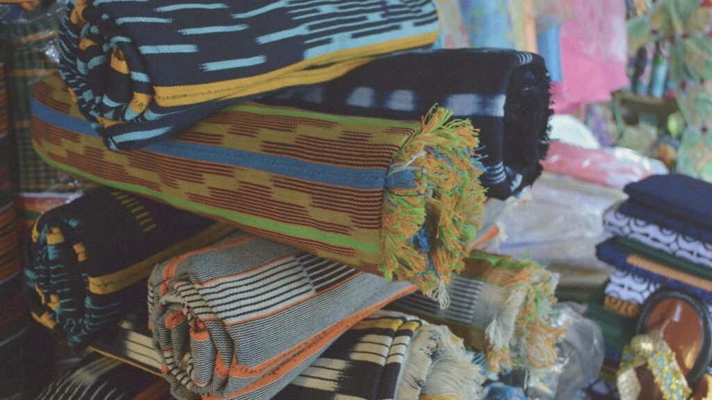 Piles de pagnes traditionnels baoulé aux motifs colorés exposés sur un marché en Côte d’Ivoire, reflets du savoir-faire artisanal akan