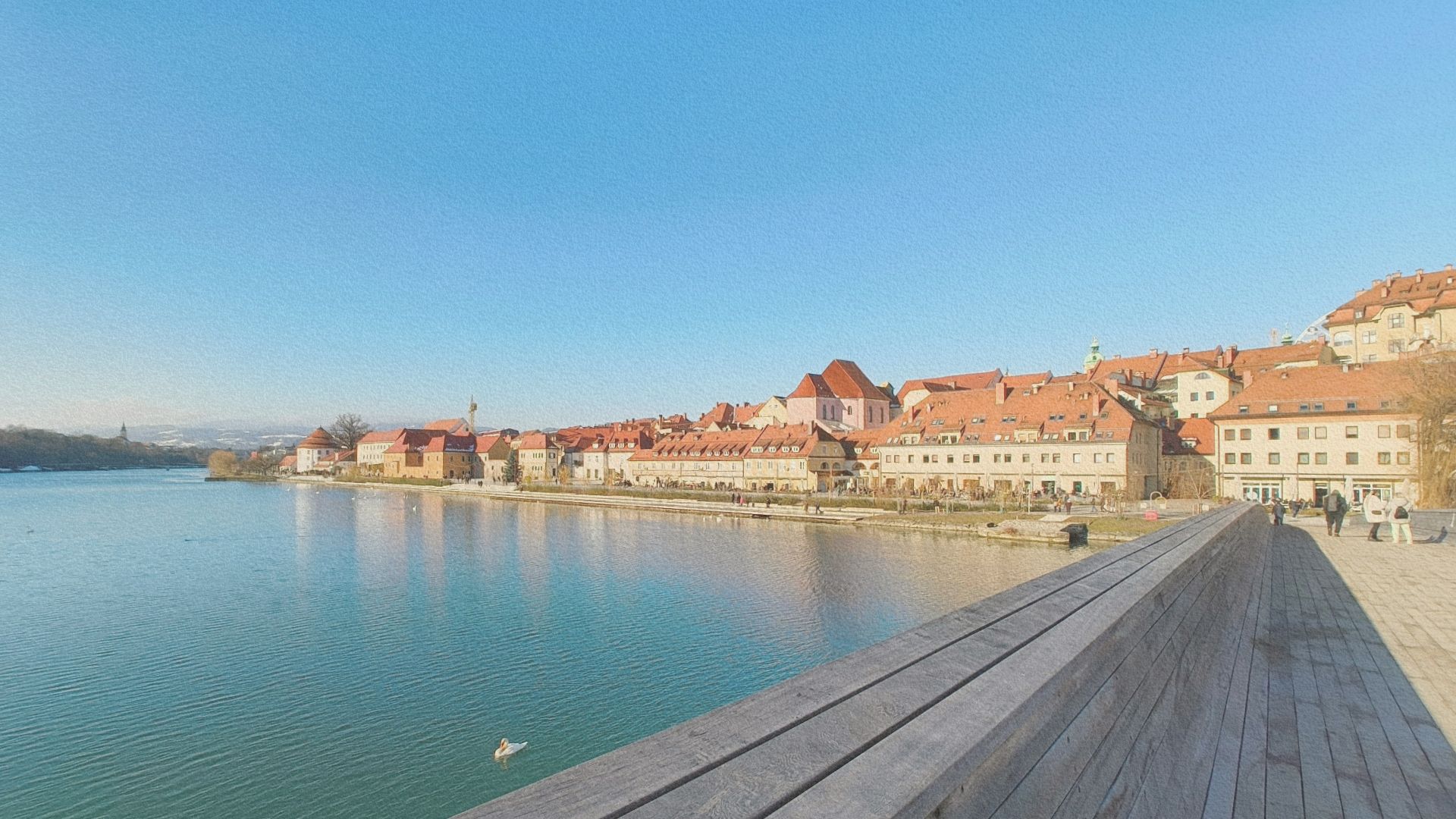 Vue sur le centre historique de Celje depuis un pont piéton, avec les toits orangés de la vieille ville se reflétant dans la rivière sous un ciel dégagé.