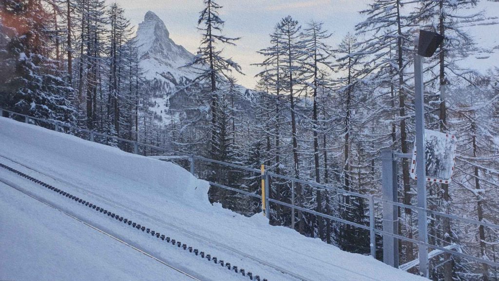 Train du Gornergrat traversant une forêt enneigée avec le Cervin en arrière-plan, révélant l’adaptation des infrastructures alpines au relief