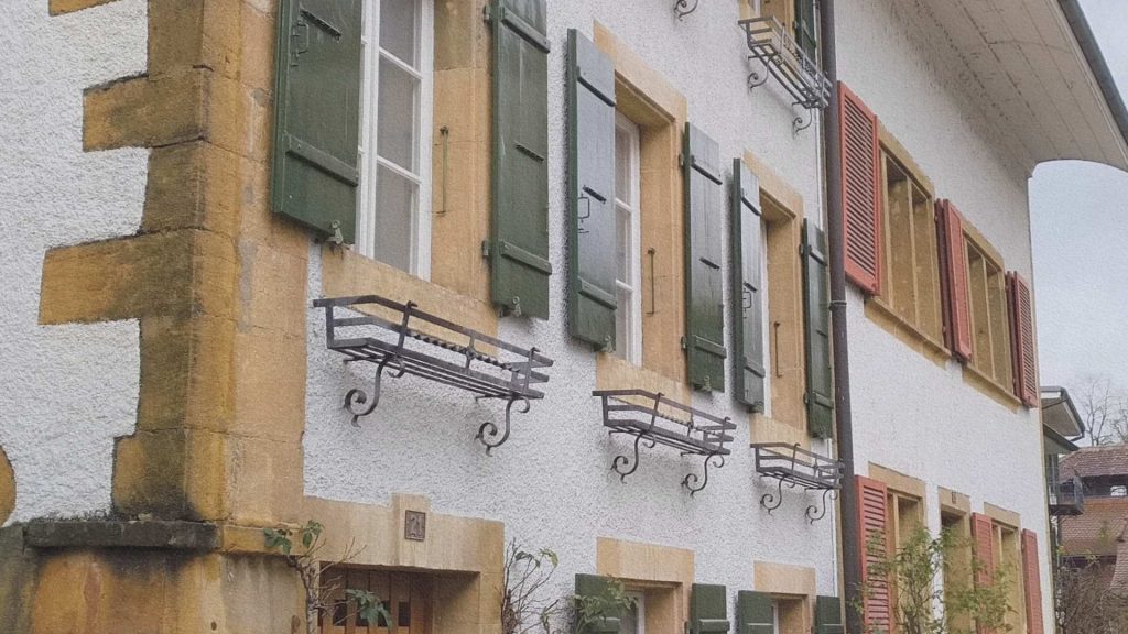 Façade traditionnelle à Morat en Suisse avec volets verts, encadrements en pierre et supports de jardinières en fer forgé