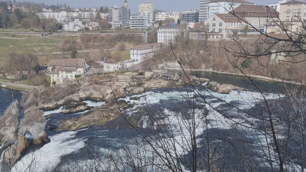 Chutes du Rhin à Neuhausen am Rheinfall en Suisse vues depuis la rive, avec bâtiments, rivière et formation rocheuse révélant la transition entre ville et force naturelle