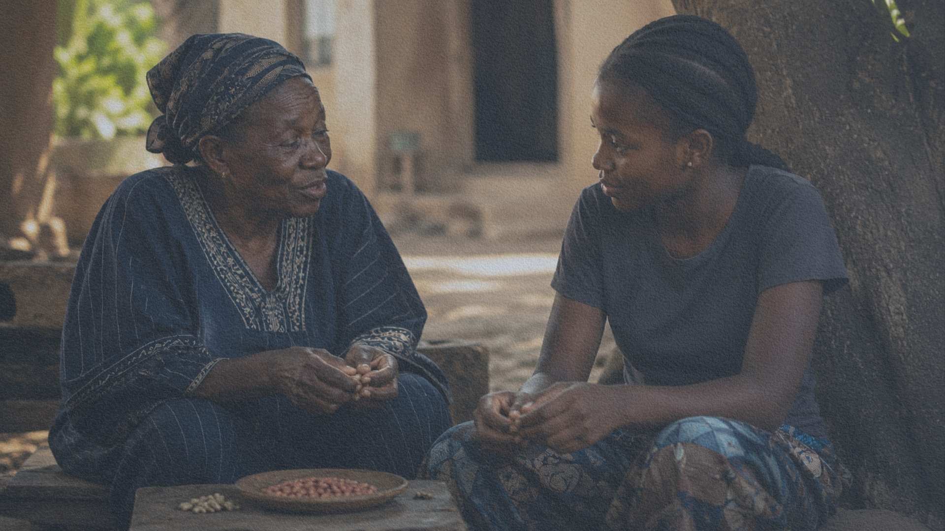 Une femme baoulé âgée partage un moment de transmission avec sa petite-fille dans une cour familiale en Côte d'Ivoire.