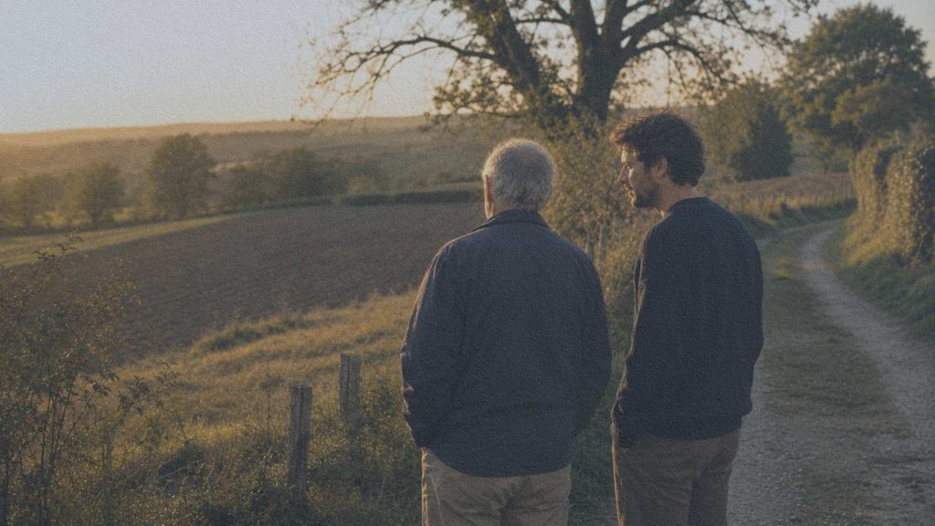 Un homme âgé et un jeune adulte observent les champs depuis un chemin rural bordé de haies à la fin de la journée.
