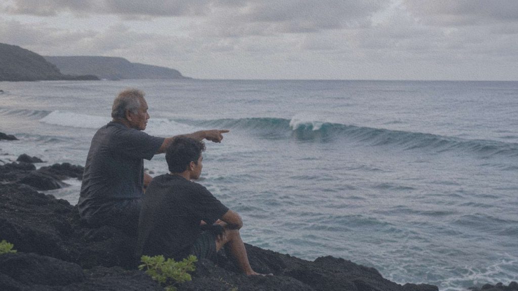 Un ancien navigateur et un jeune apprenti observent une vague depuis des rochers volcaniques, face au Pacifique.