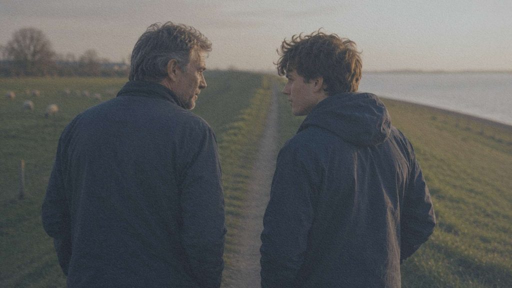Un homme âgé et un jeune adulte échangent quelques mots sur une digue néerlandaise au coucher du soleil, devant un paysage de prairies et de moutons.