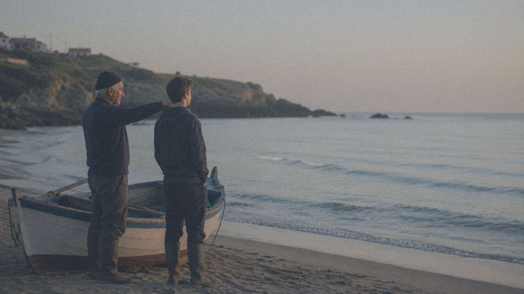 Un vieux pêcheur portugais montre discrètement une série de petites vagues à un jeune apprenti près d'une barque traditionnelle posée sur le rivage.
