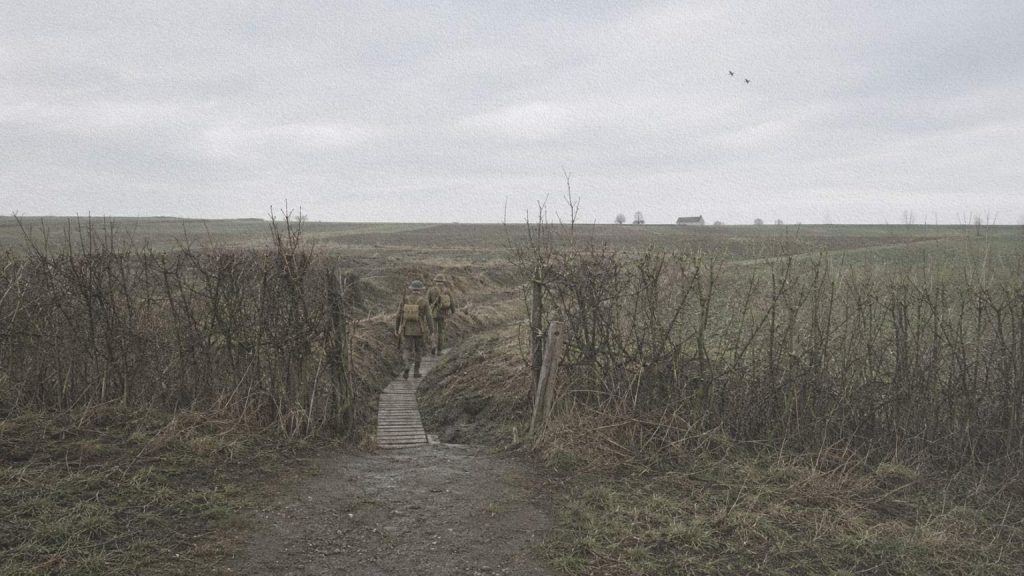 Brèche dans une haie donnant accès à des caillebotis et à des tranchées dissimulées dans un paysage rural du nord de la France.