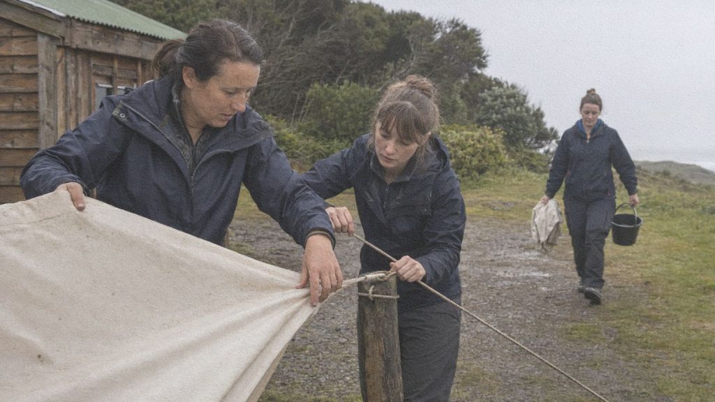 Trois femmes réparent une toile endommagée par le vent dans un camping côtier après la pluie.